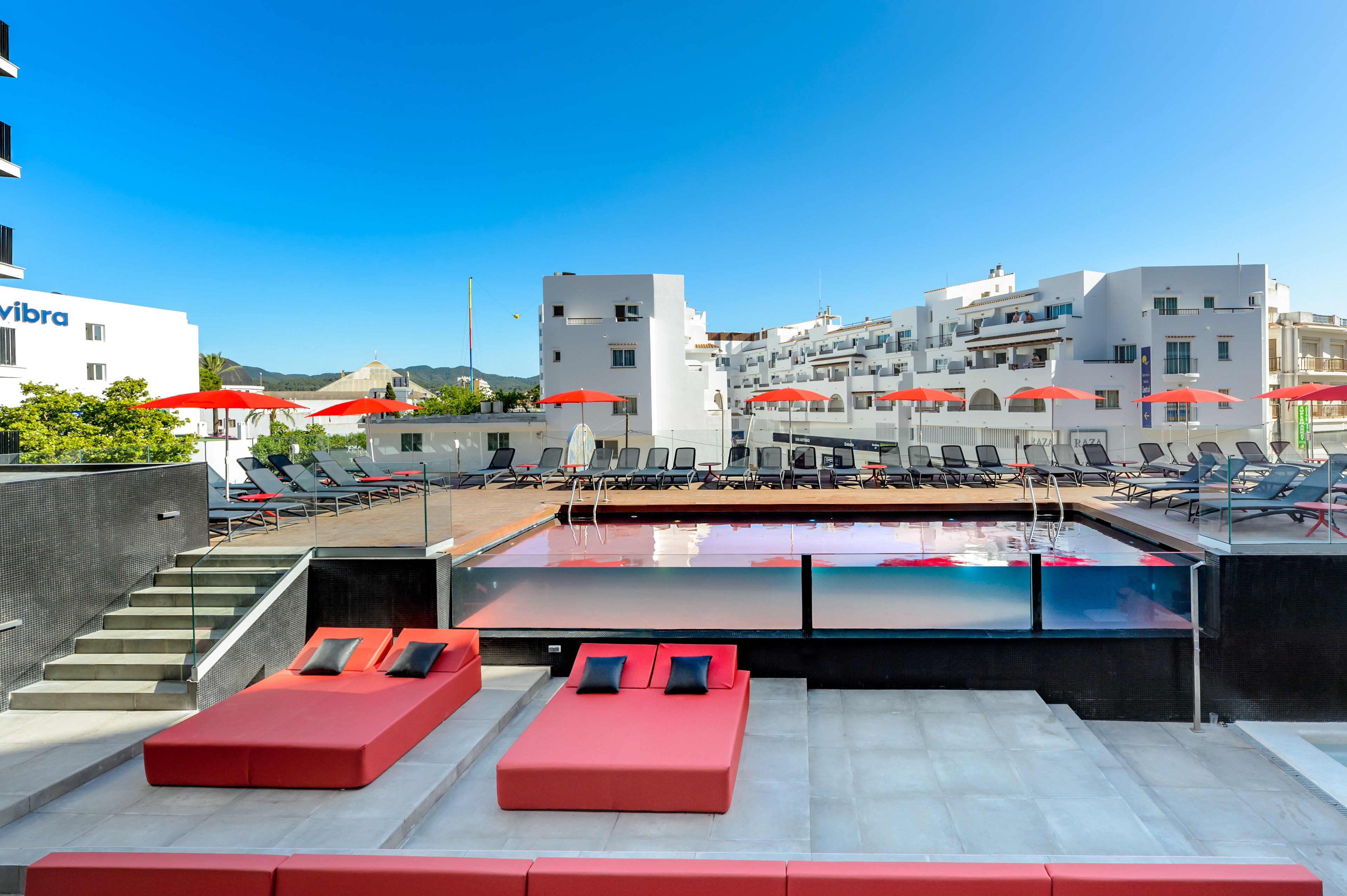 A rooftop pool area with red lounge chairs and umbrellas, a clear blue sky, and white buildings in the background.