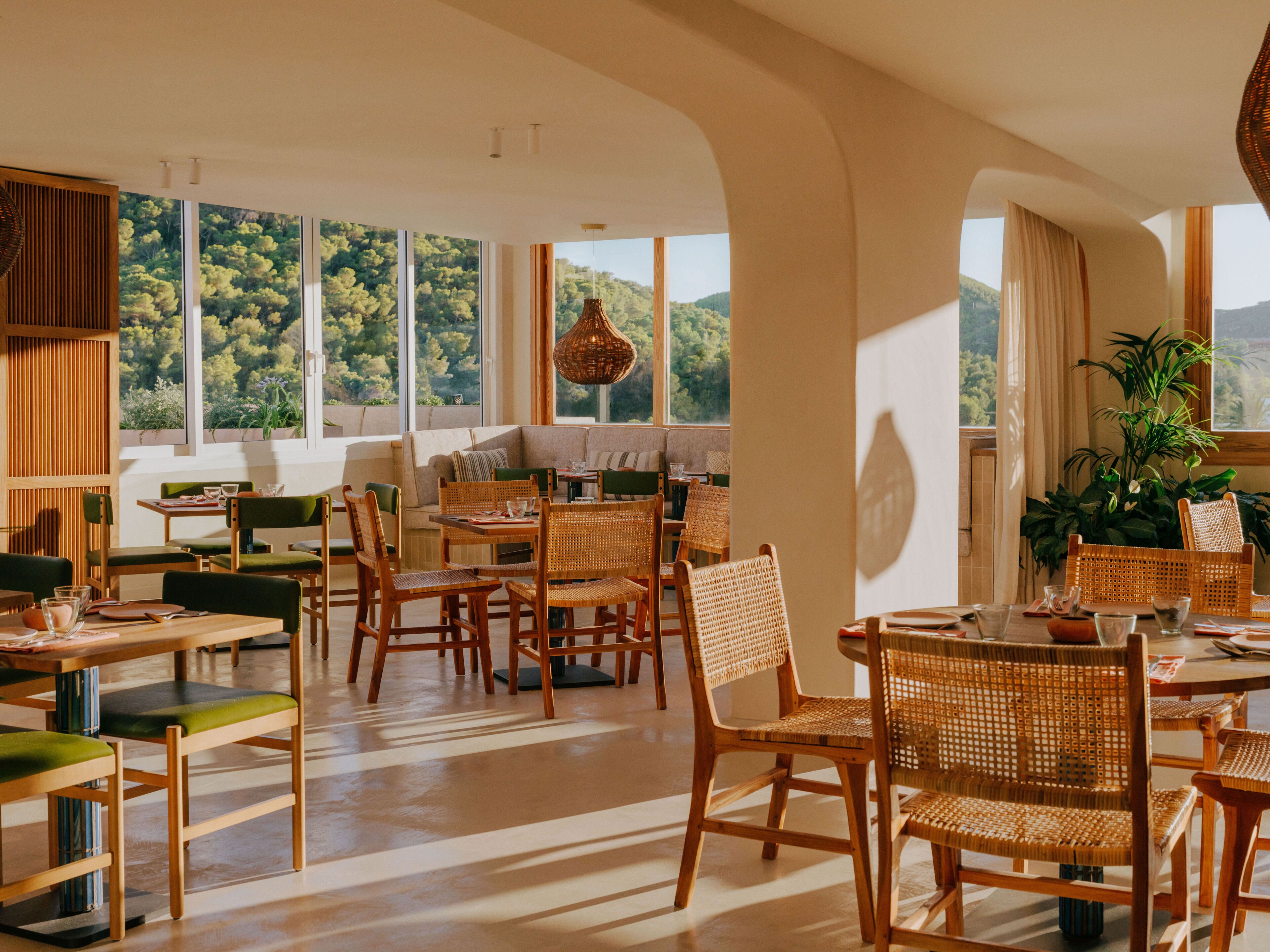 A spacious dining area with high ceilings, large windows offering a view of greenery, and a mix of wooden and woven furniture.