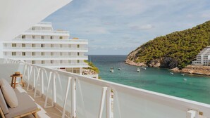 Un balcon avec vue sur la mer, un bâtiment blanc et une colline boisée.