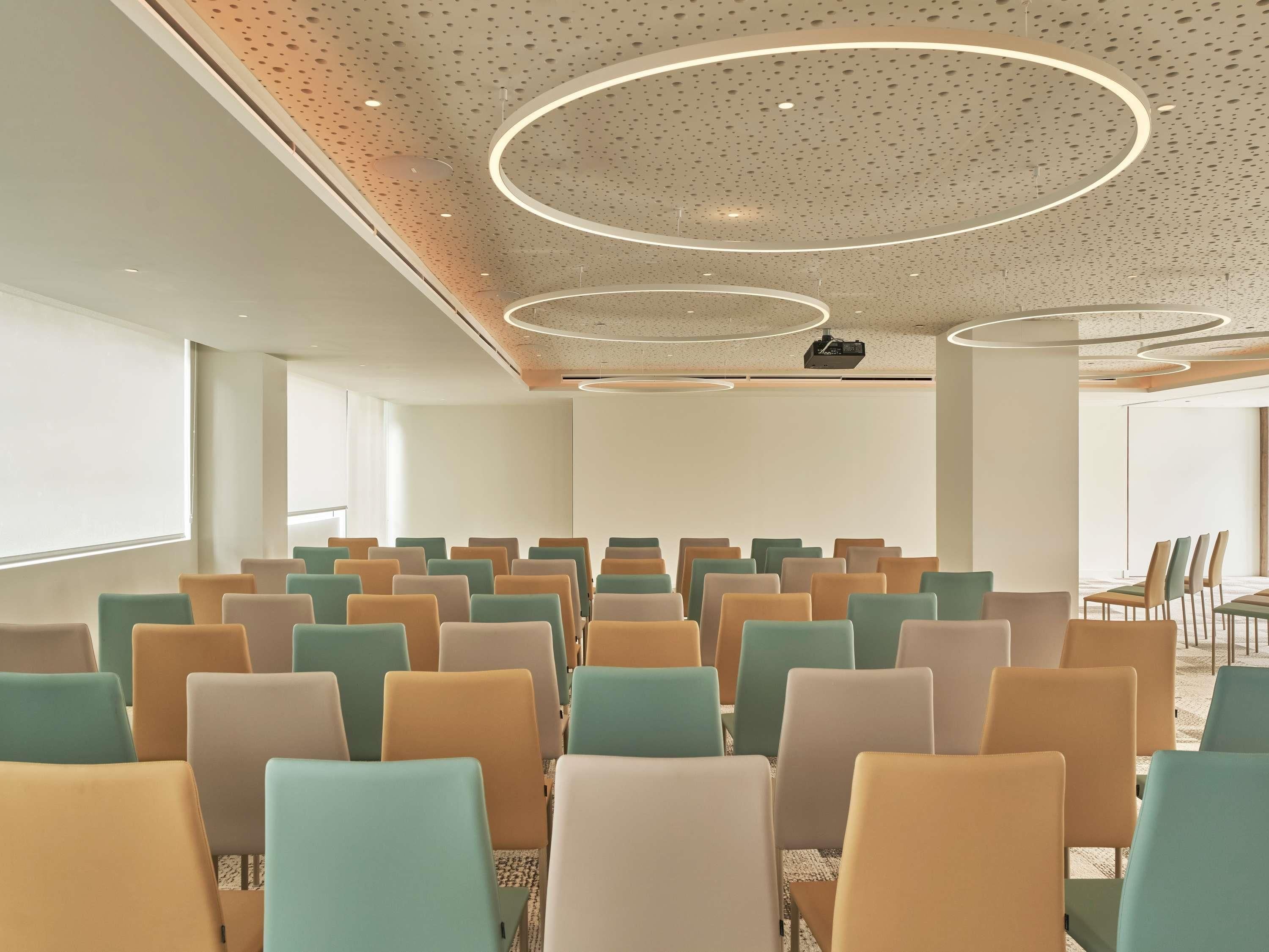 Une salle de conférence avec des rangées de chaises, un écran de projection et un plafond orné de luminaires circulaires.