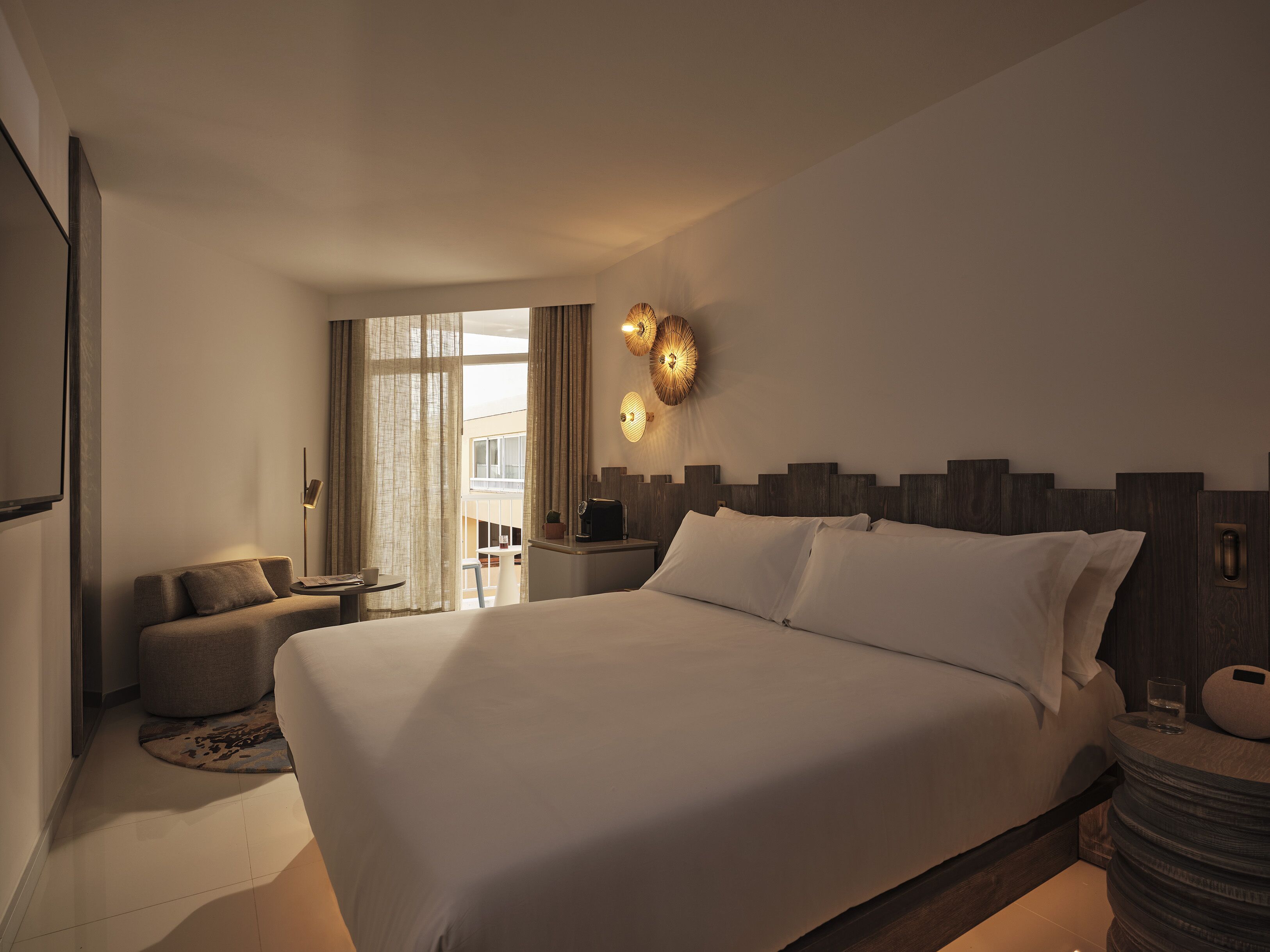 A modern hotel room with a large bed, a sofa, a small table, and a view of the outside.