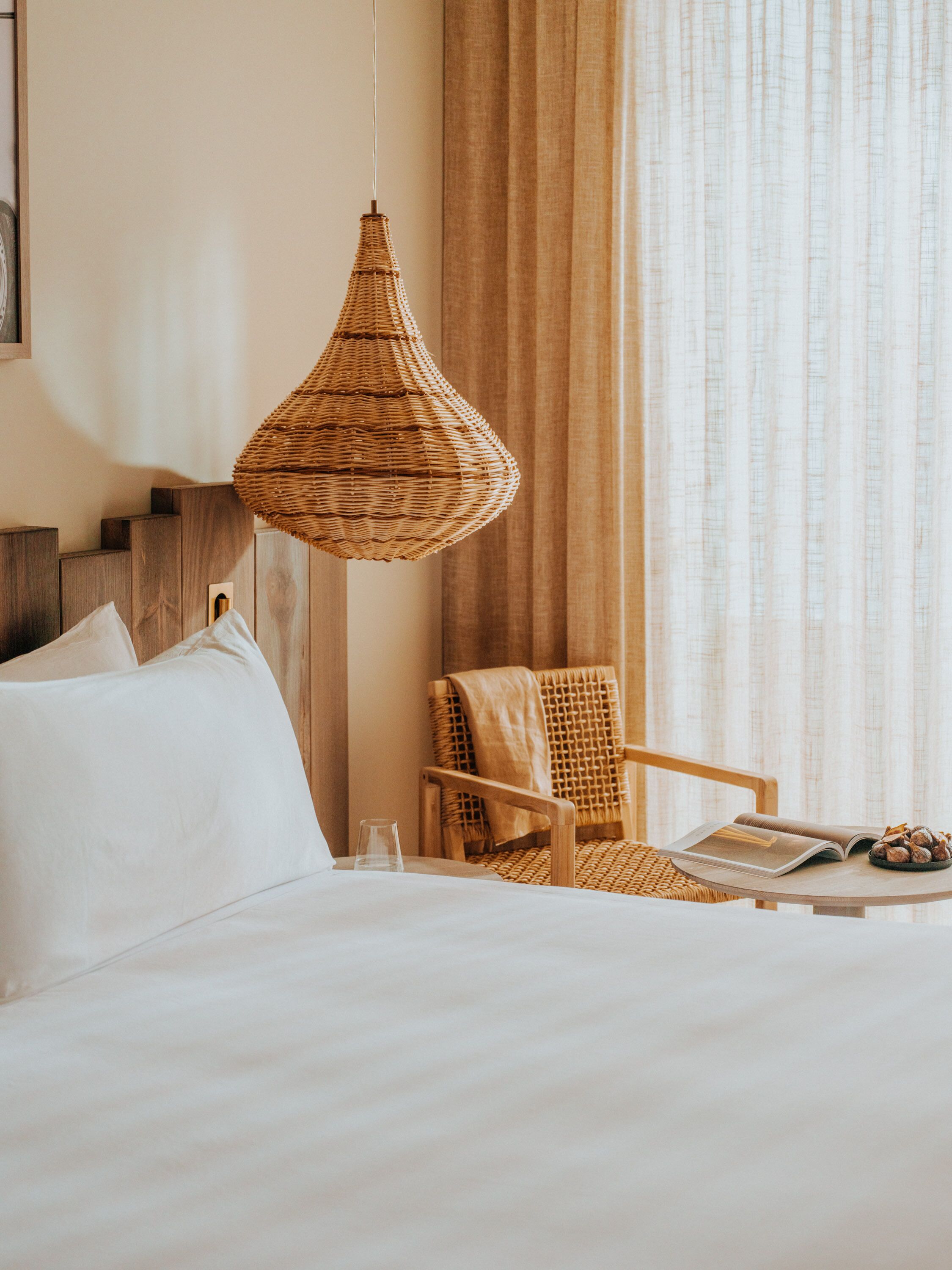 Una habitación de hotel con una cama, una silla de mimbre, una mesa con un libro y un plato de nueces, y una lámpara colgante.