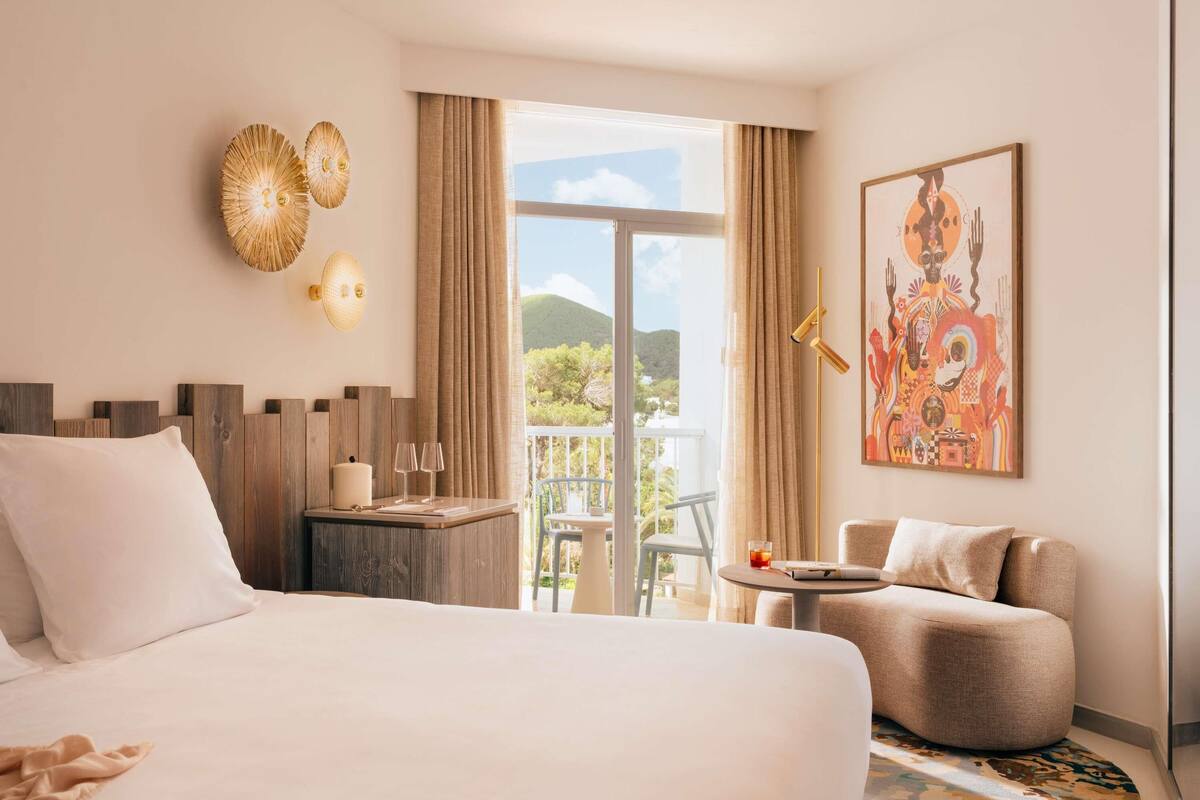 Une chambre d’hôtel avec un lit, une petite table avec des verres, une chaise, un balcon avec vue et un tableau au mur.