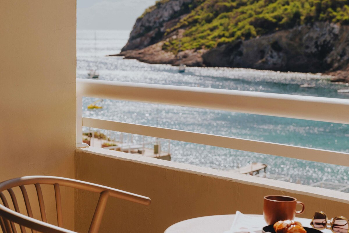 Un balcón con vista al mar, una silla, una mesa con una taza y algunos objetos sobre la mesa.