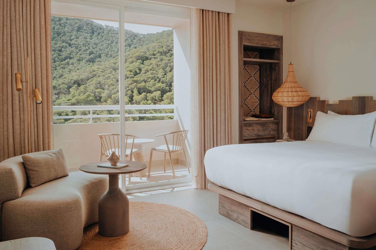Une chambre d’hôtel avec un grand lit, une petite table ronde, deux chaises, un canapé, une table de chevet en bois, une lampe en osier et une vue sur une colline boisée.
