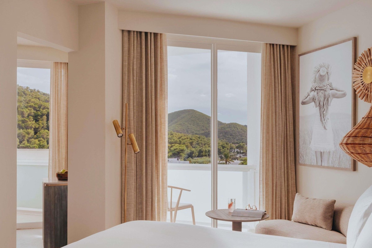 Habitación de hotel con una cama grande, una mesita con una silla y vistas a un lago y montañas a través de ventanas de techo a piso.