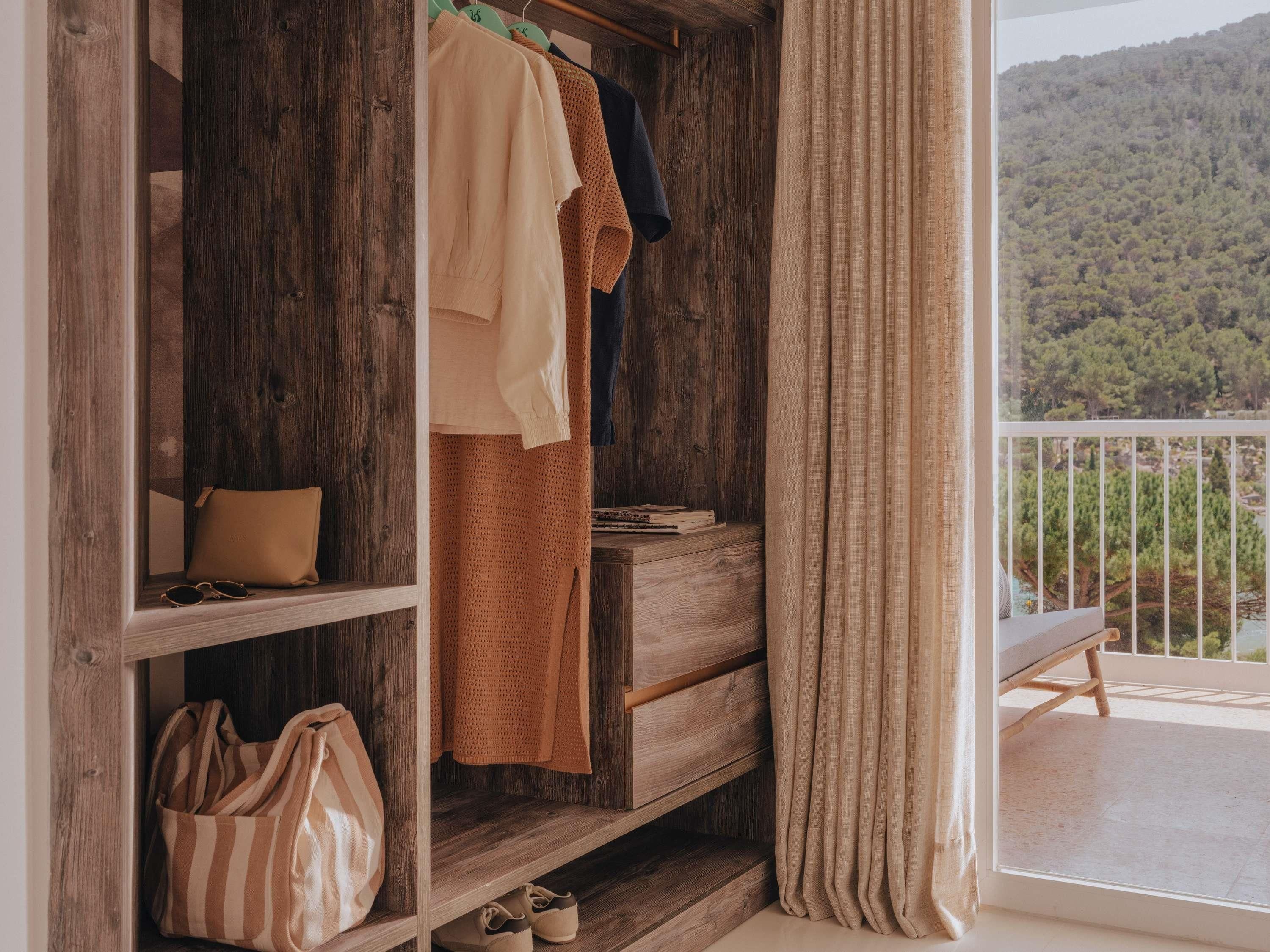 A wooden wardrobe with hanging clothes, a shelf with a bag and shoes, and a view of a forested hillside through a balcony.