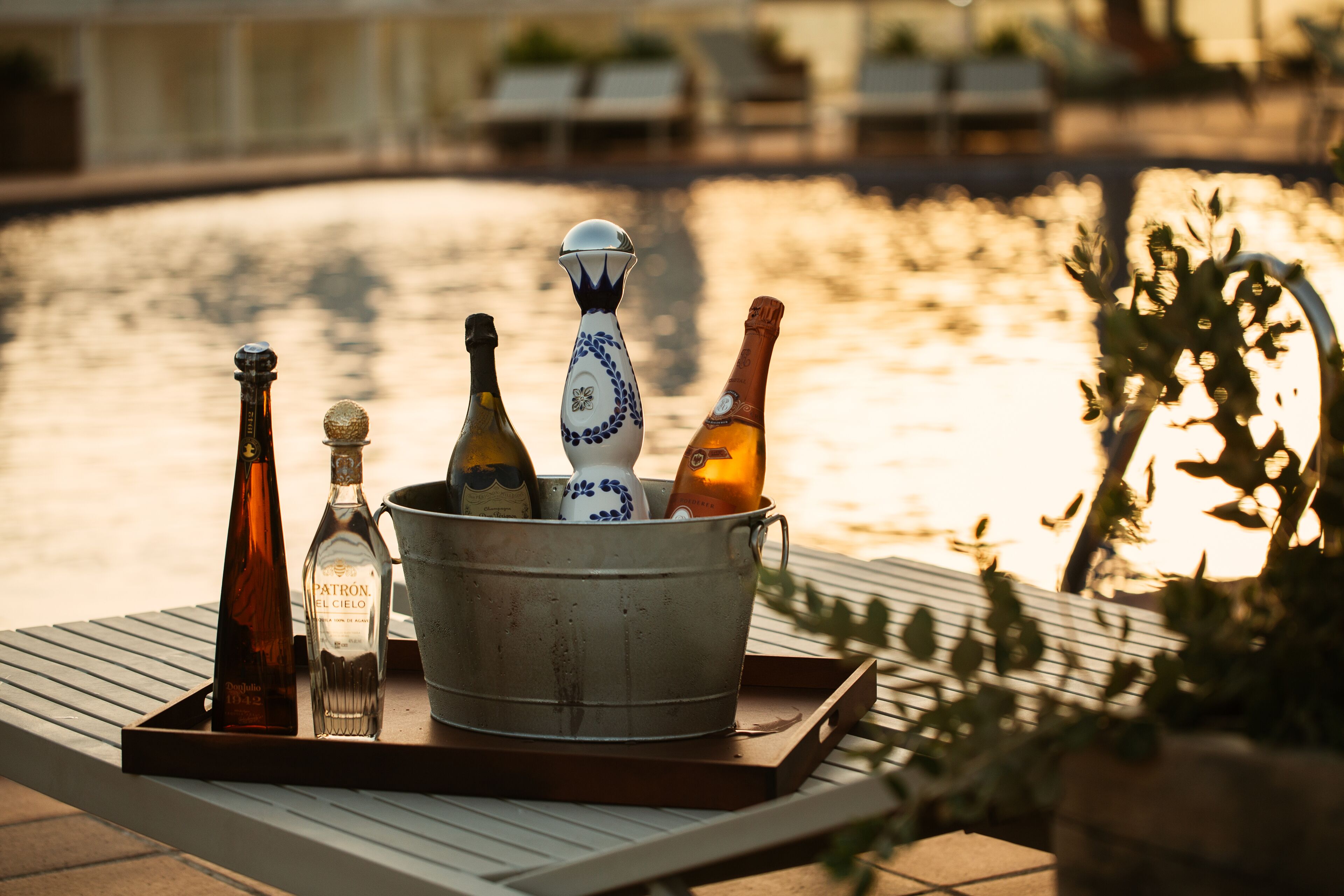 A metal bucket filled with bottles on a tray, placed on a table with a view of a body of water and lounge chairs in the background.