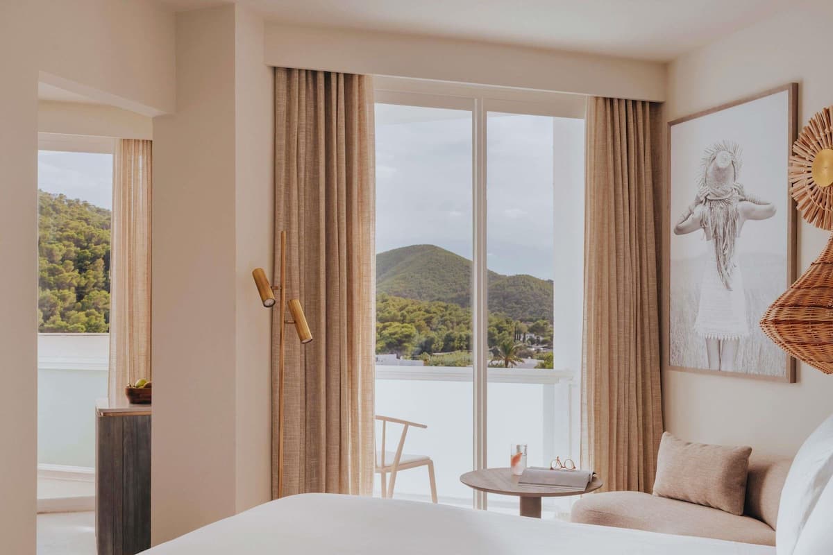 Une chambre d’hôtel avec un grand lit, une petite table avec une chaise, et une vue sur un lac et des montagnes grâce à de grandes fenêtres du sol au plafond.