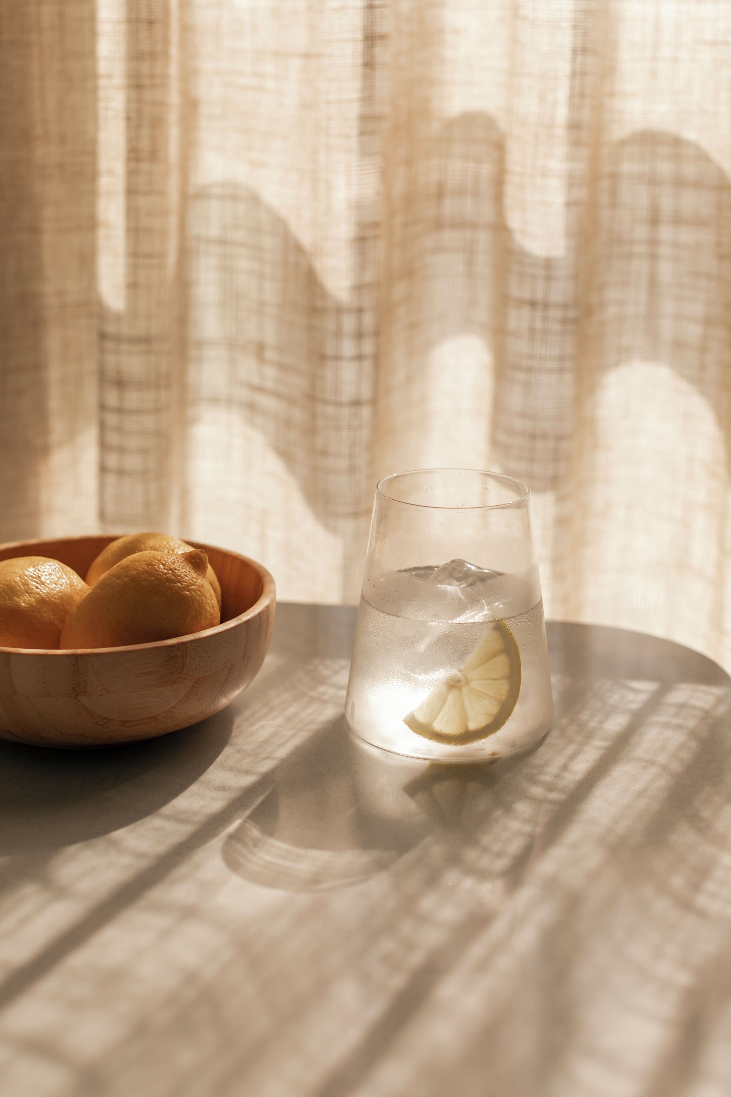 Een glas water met een plakje citroen op een tafel, met een houten kom vol citroenen op de achtergrond.
