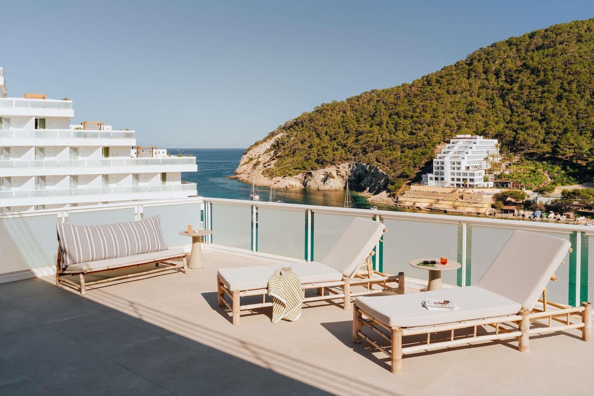 A balcony with lounge chairs and a small table, overlooking a coastal area with buildings and a hill.