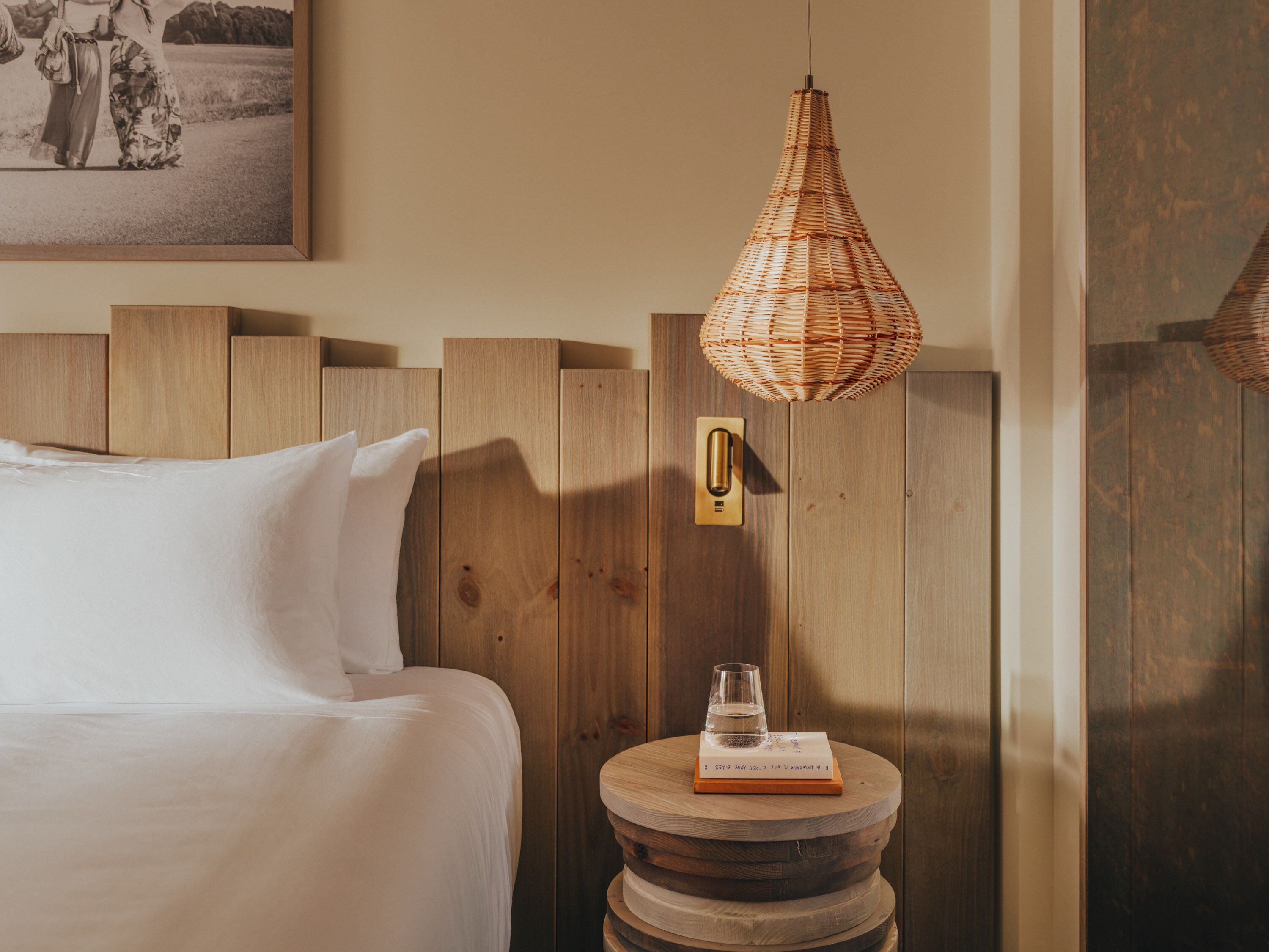 Una cama bien hecha con sábanas blancas, un cabecero de madera, una mesita de noche de madera con un vaso de agua y un libro, y una lámpara colgante de mimbre.