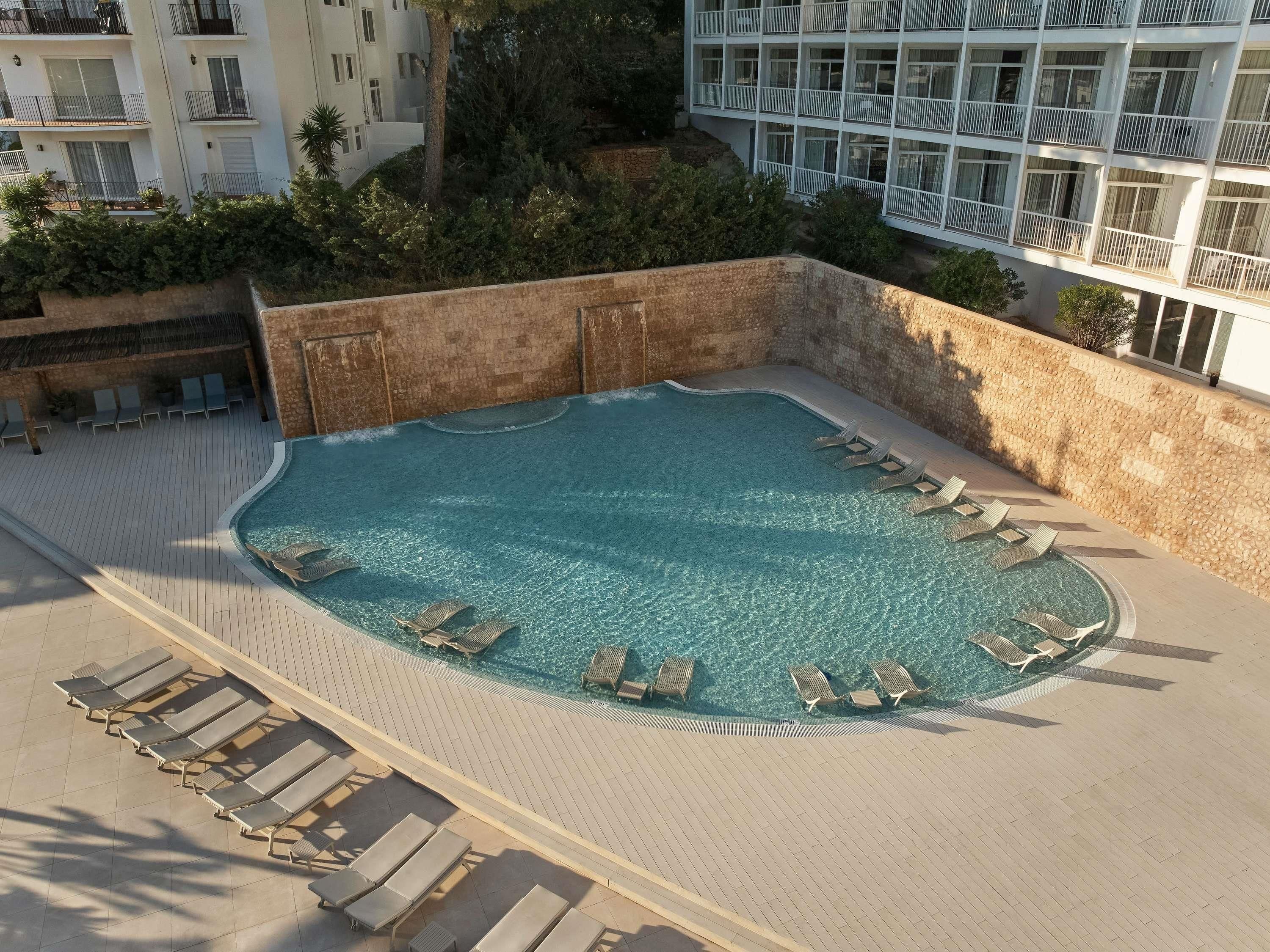A swimming pool with lounge chairs, a stone wall, and a building in the background.
