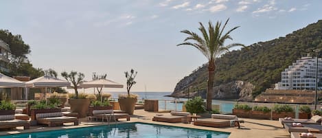 Zona de piscina con sillones de descanso y sombrillas, una piscina cristalina y vistas al mar y las colinas.