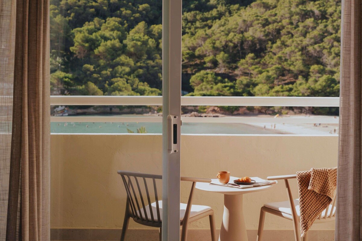 Un balcón con vistas a una playa y un bosque. Hay una mesa con una taza y un plato.