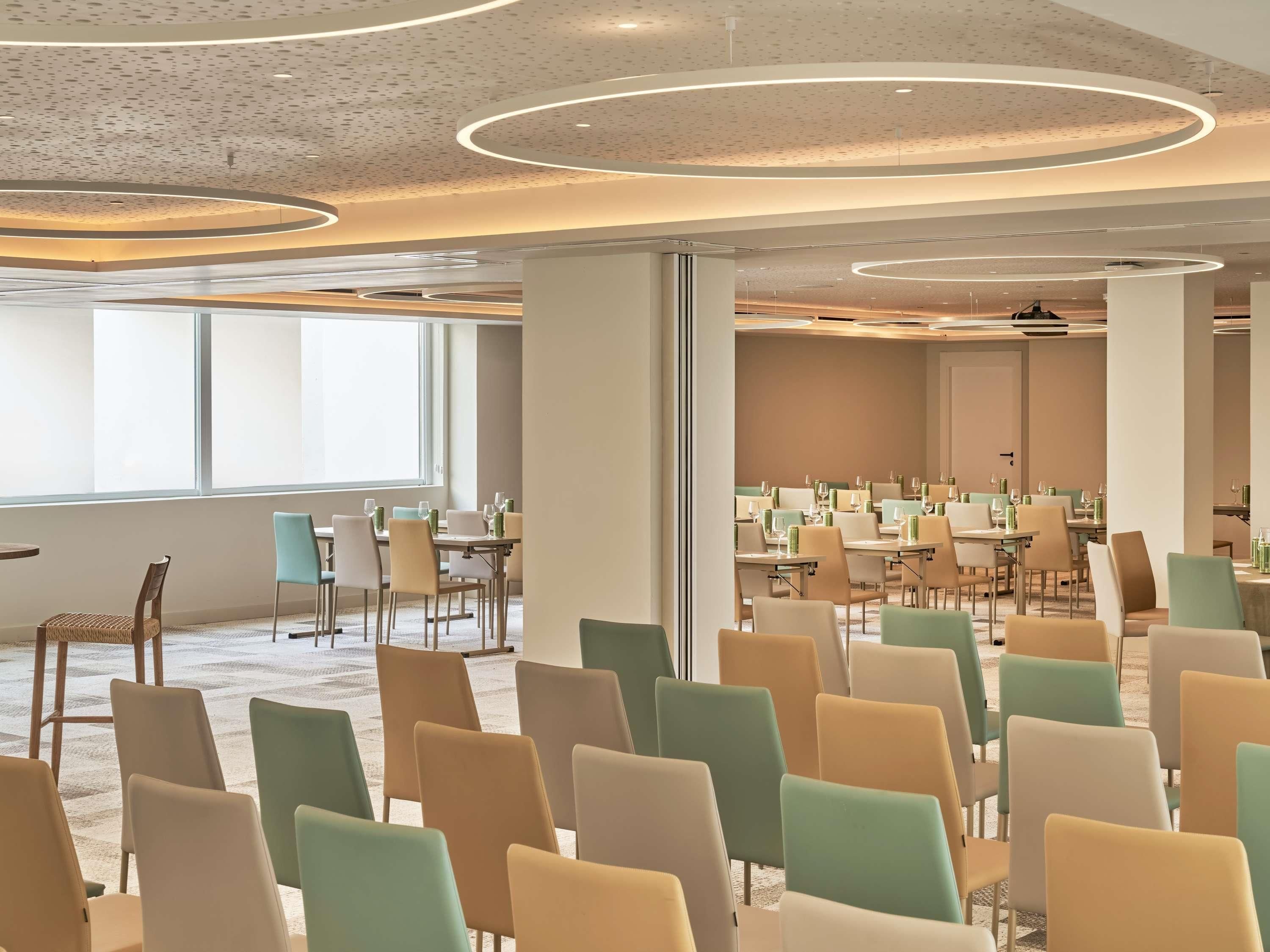 Een vergaderzaal met rijen stoelen, een groot raam en een modern plafond.