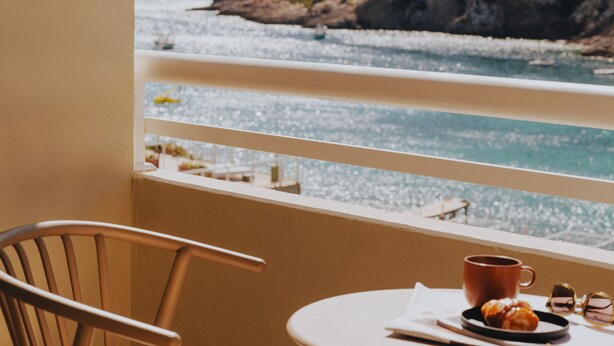 Un balcon avec vue sur la mer et les montagnes, comprenant deux chaises et une table avec une tasse et une pâtisserie.