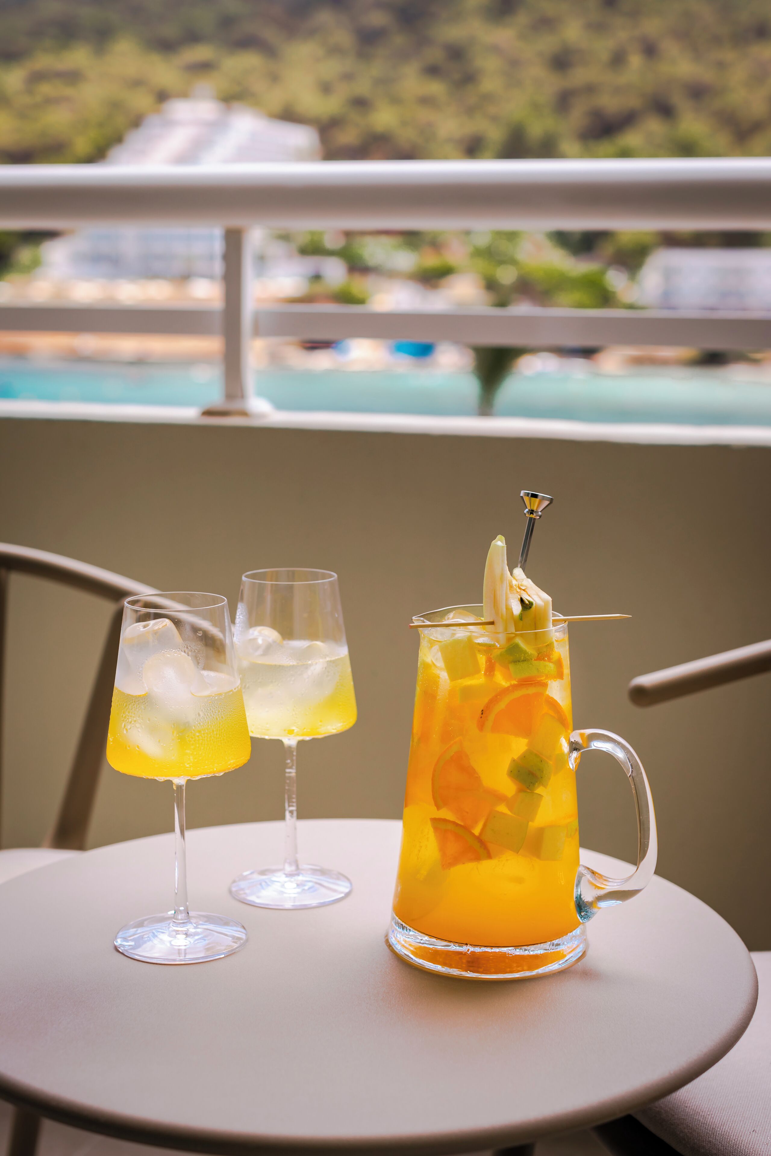 Zwei Gläser und eine Karaffe mit einem gelben Getränk, Eiswürfeln und Früchten auf einem Tisch mit Blick auf einen Pool und Grünflächen.