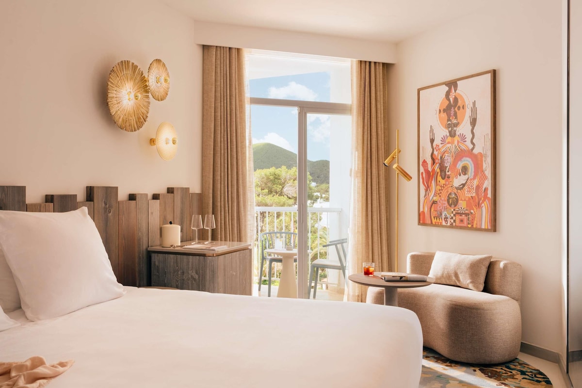 Una habitación de hotel con una cama, una mesita con vasos, una silla, un balcón con vistas y un cuadro en la pared.