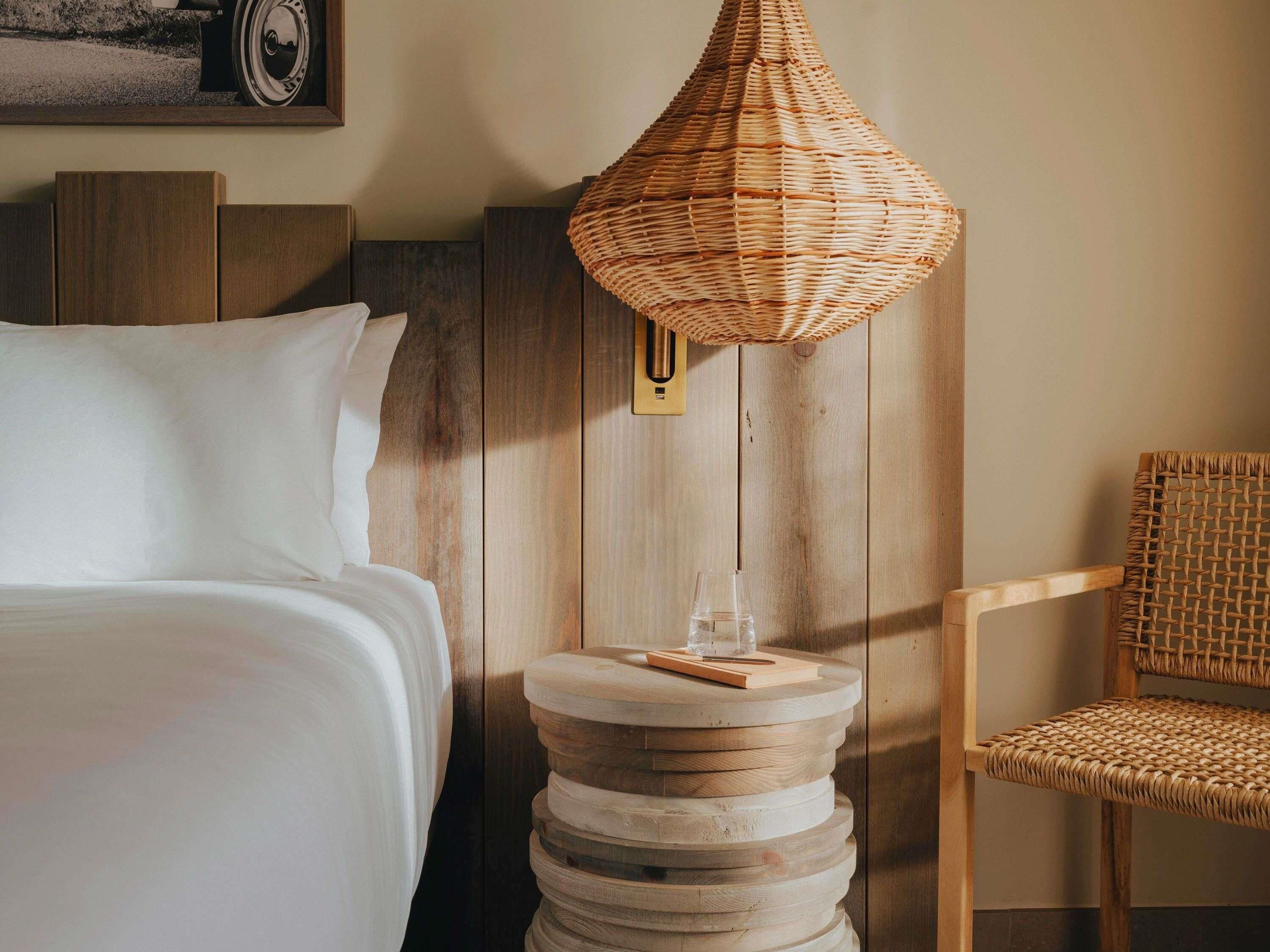 A bed with white linens, a round wooden side table with a glass of water and a book, a wicker chair, and a wicker lamp on a wooden wall.