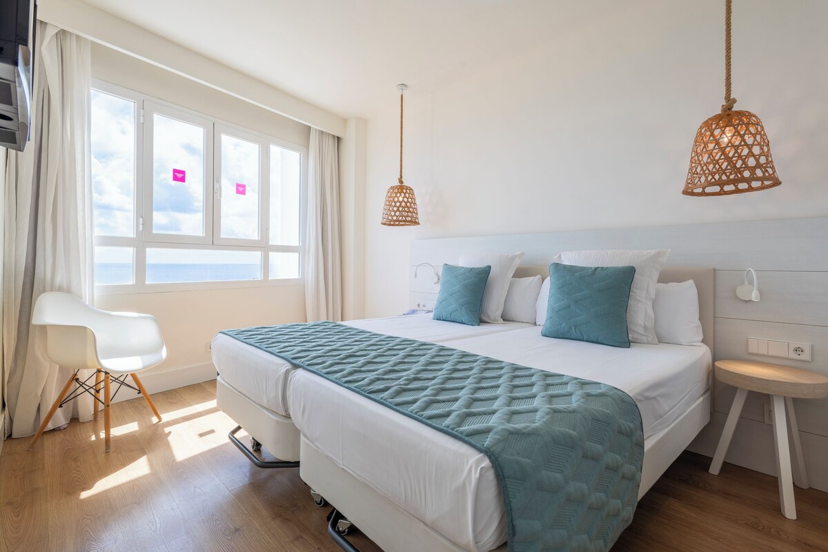 A modern hotel room with a large bed, a white chair, and a view of the sea through the windows.