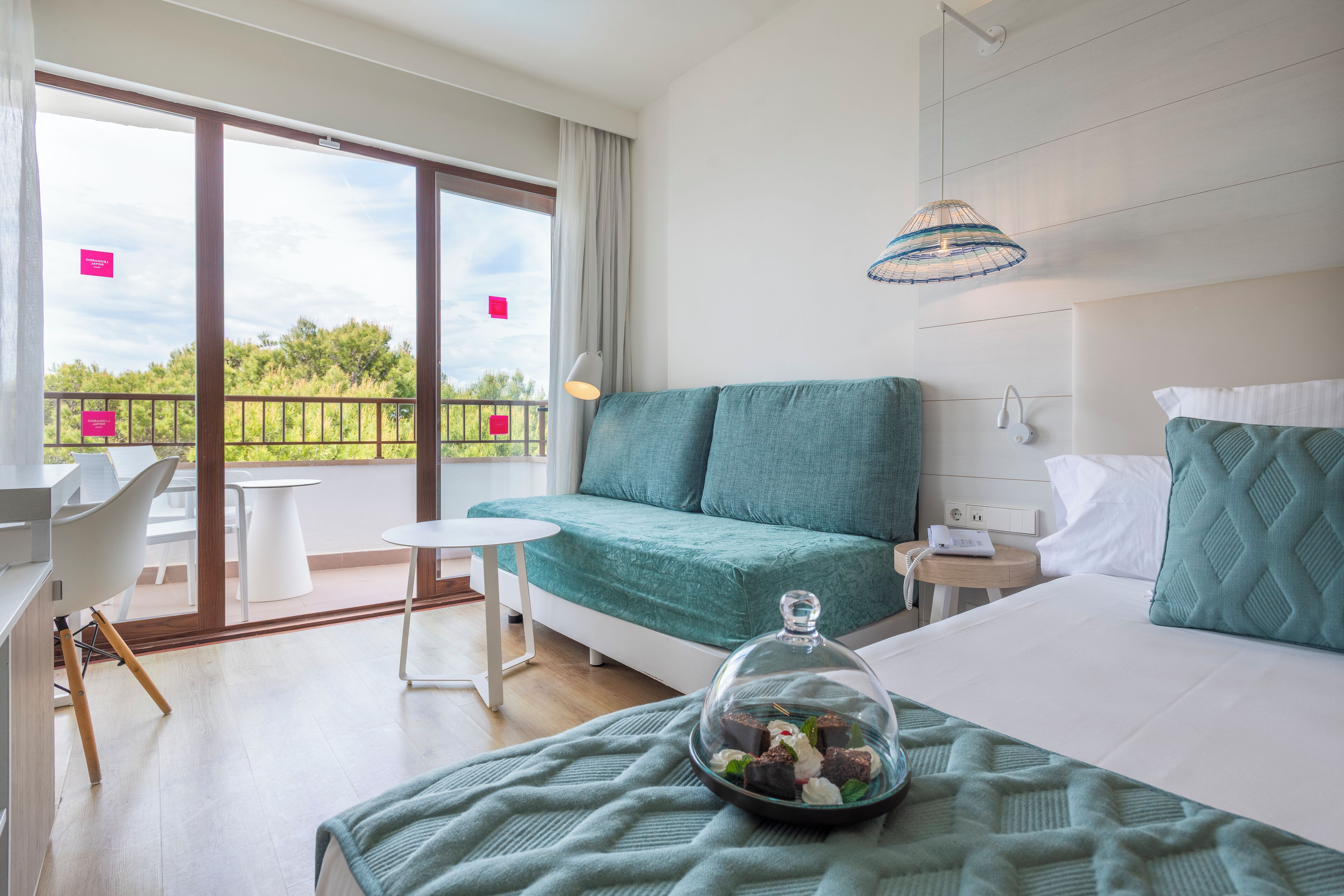 A modern hotel room with a balcony, a bed with a glass dome, a small table, and a sofa.