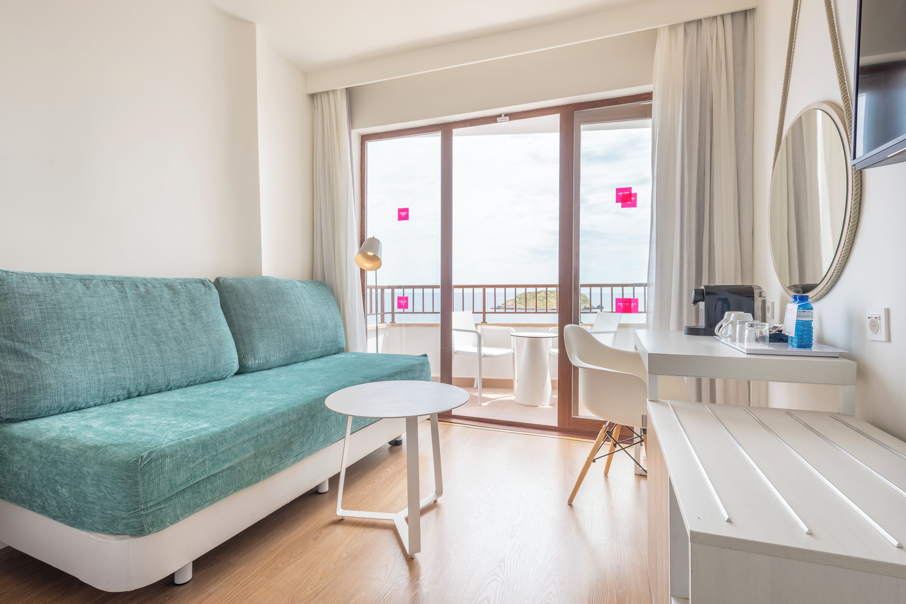 Habitación de hotel moderna con sofá azul aguamarina, una mesita redonda pequeña, un escritorio blanco con espejo y un balcón con vistas.