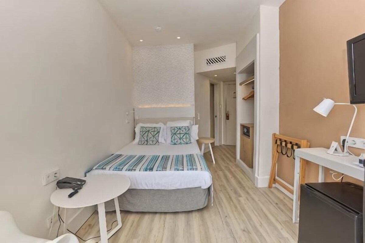 A compact hotel room with a bed, bedside table, chair, desk, and a small kitchenette.
