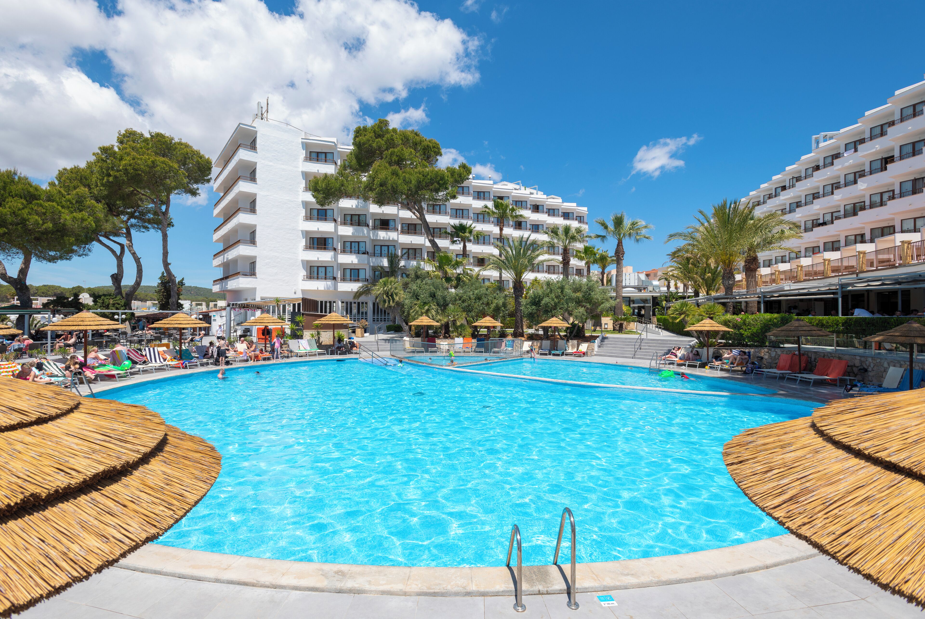 A large outdoor swimming pool surrounded by palm trees and lounge chairs, with a multi-story hotel building in the background.