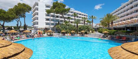 A large outdoor swimming pool surrounded by palm trees and lounge chairs, with a multi-story hotel building in the background.