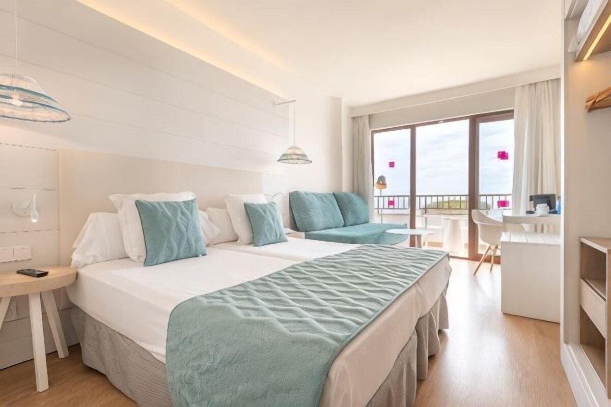 A hotel room with two beds, a desk, a chair, and a balcony with a view.