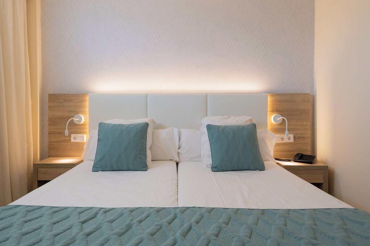 A double bed with a quilted bedspread, two pillows, bedside tables with lamps, and a wooden headboard.