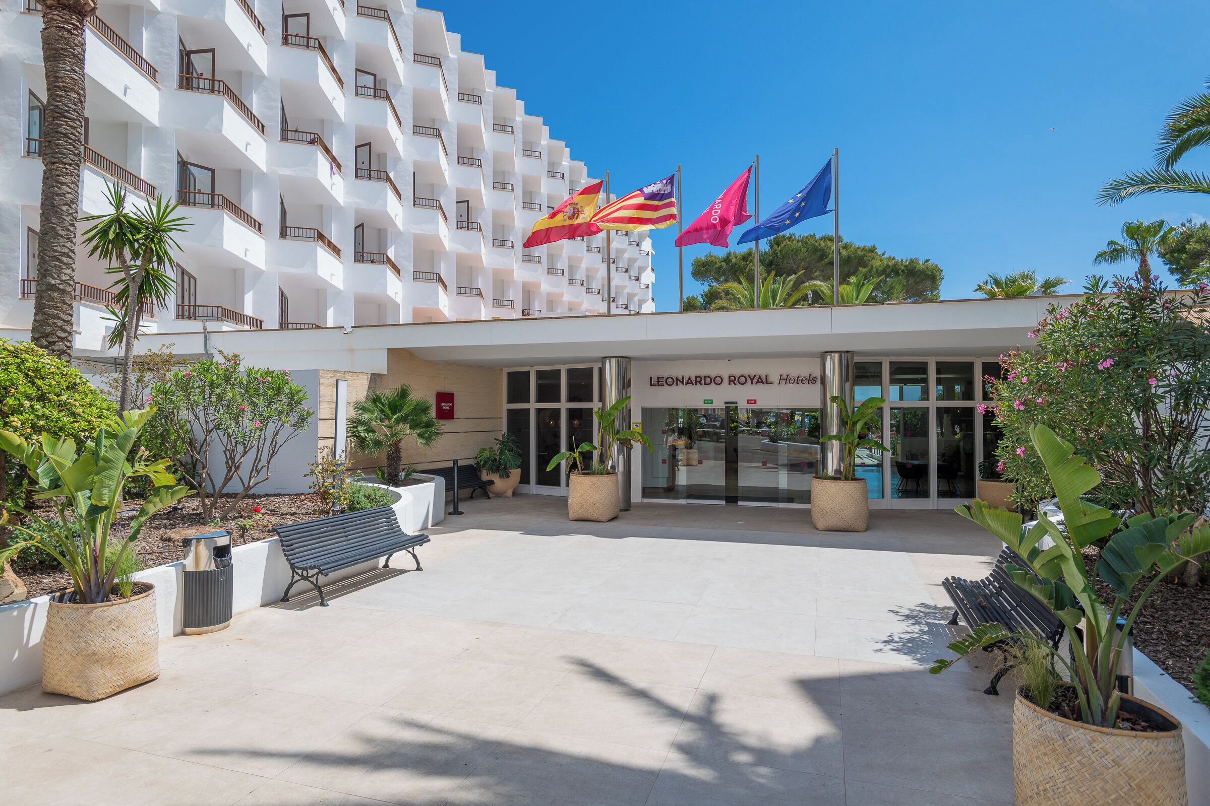 Entrada de hotel com fachada branca, várias varandas e bandeiras, incluindo a bandeira espanhola e a bandeira da União Europeia.