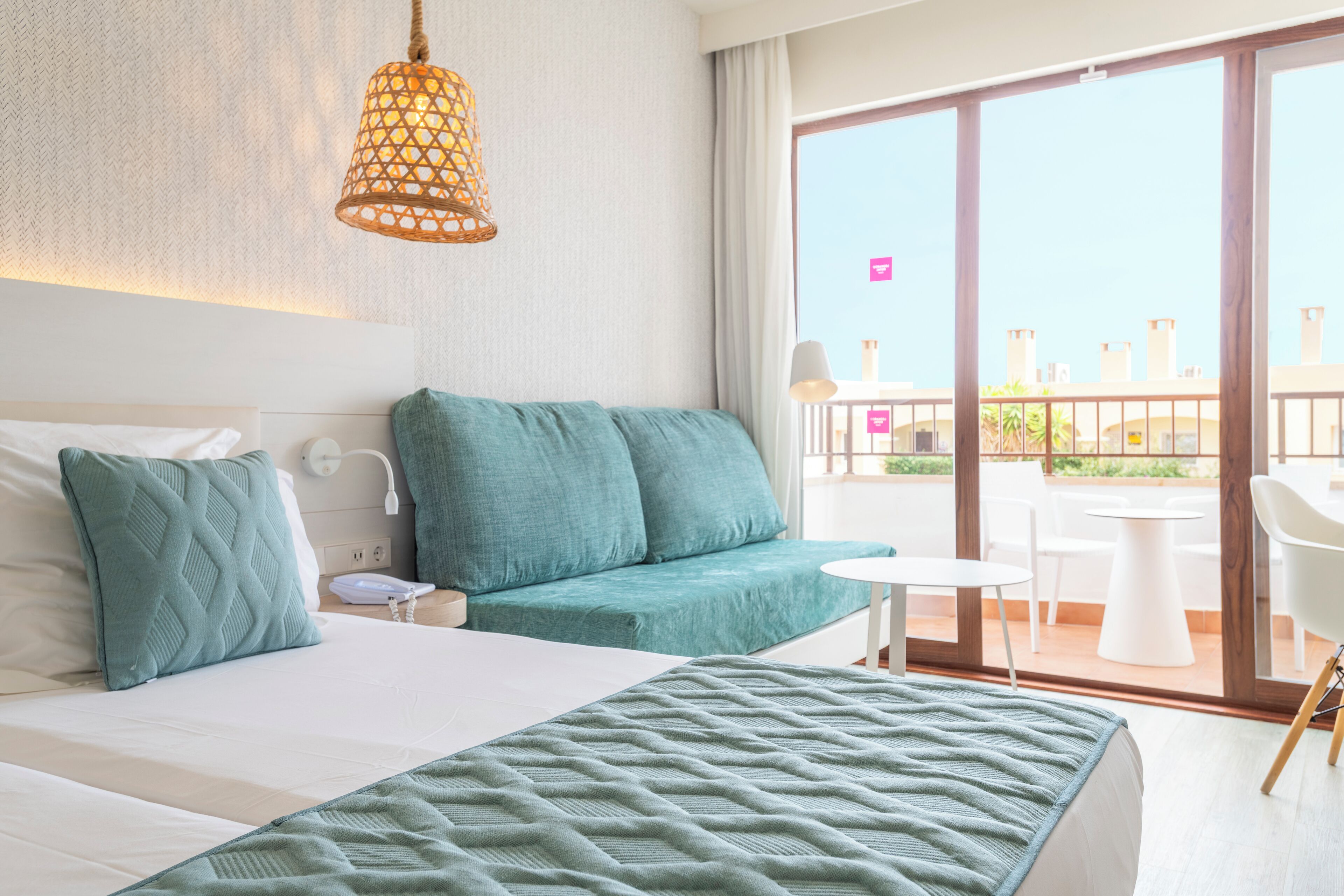 A hotel room with a bed, a sofa, a small table, and a balcony with outdoor seating.