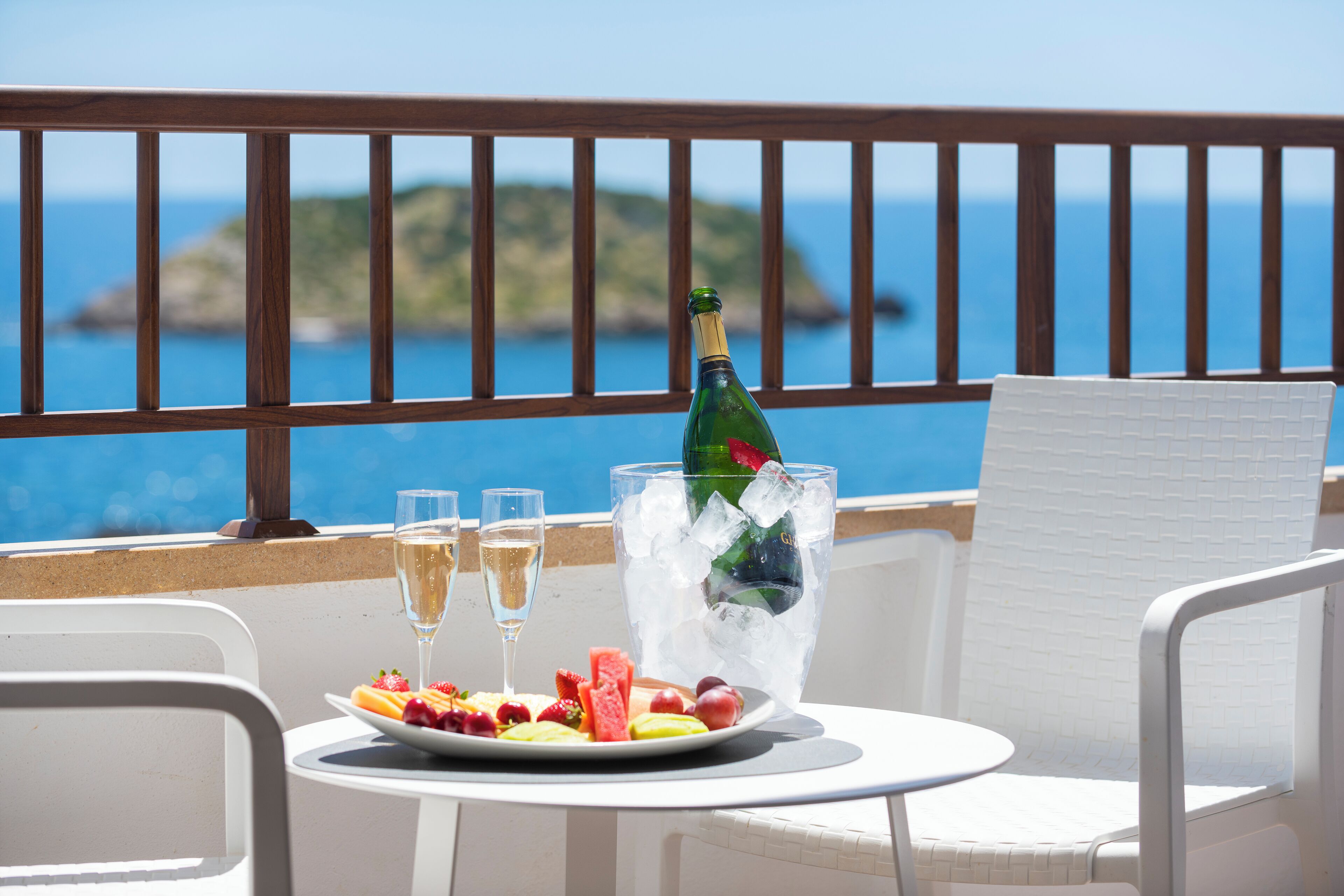 A table with a bottle of champagne, glasses, and a plate of fruit on a balcony with a view of the sea.
