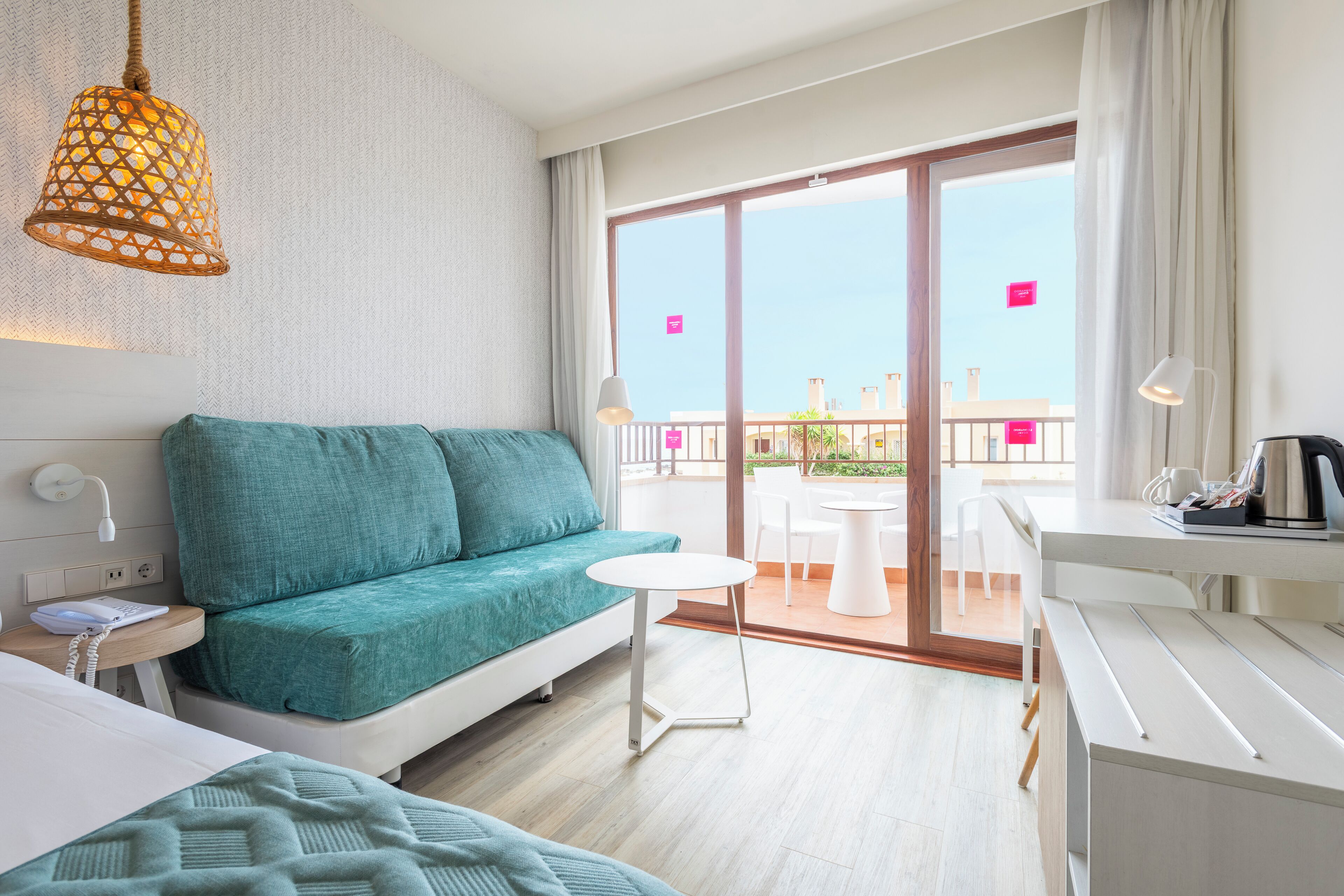 Una habitación de hotel moderna con un sofá color turquesa, una pequeña mesa redonda, un escritorio con lámpara y un balcón con vista a la ciudad.
