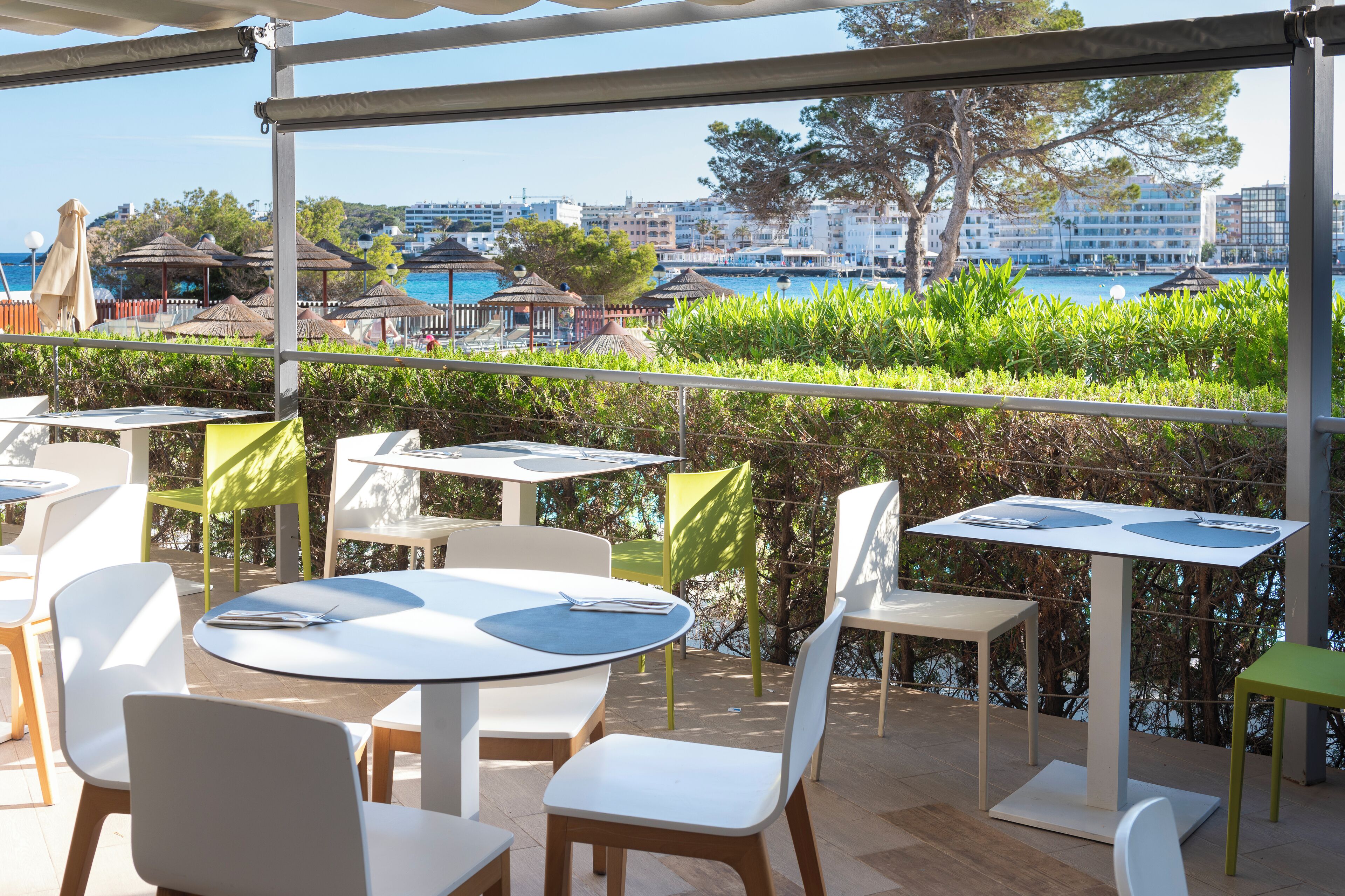 Une terrasse extérieure avec des tables et des chaises, offrant une vue sur un plan d’eau et des bâtiments.