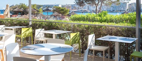 Une terrasse extérieure avec des tables et des chaises, offrant une vue sur un plan d’eau et des bâtiments.