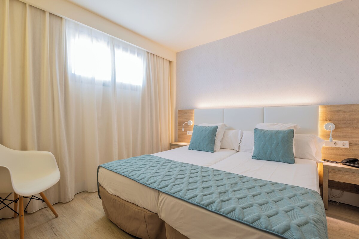 A hotel room with a large bed, two pillows, a bedside table with a lamp, and a chair.