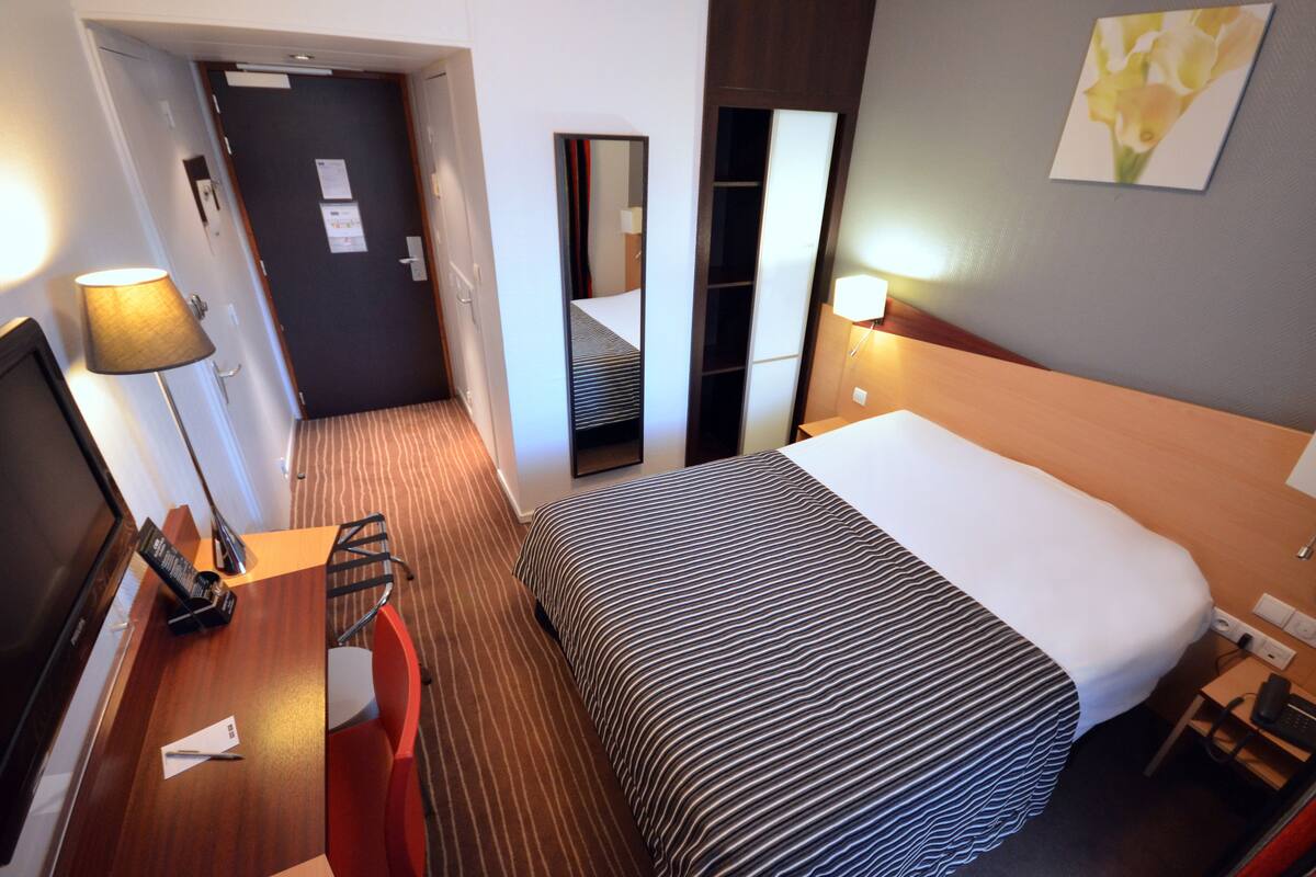 Une chambre d’hôtel avec un lit, un bureau doté d’une lampe, une chaise, un miroir et un tableau accroché au mur.