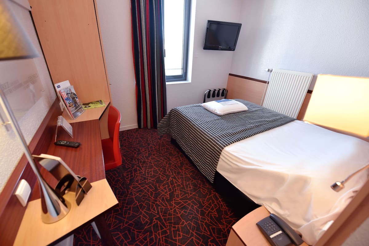 Une chambre d’hôtel avec un lit, un bureau avec une lampe, un téléviseur fixé au mur et une fenêtre avec des rideaux.