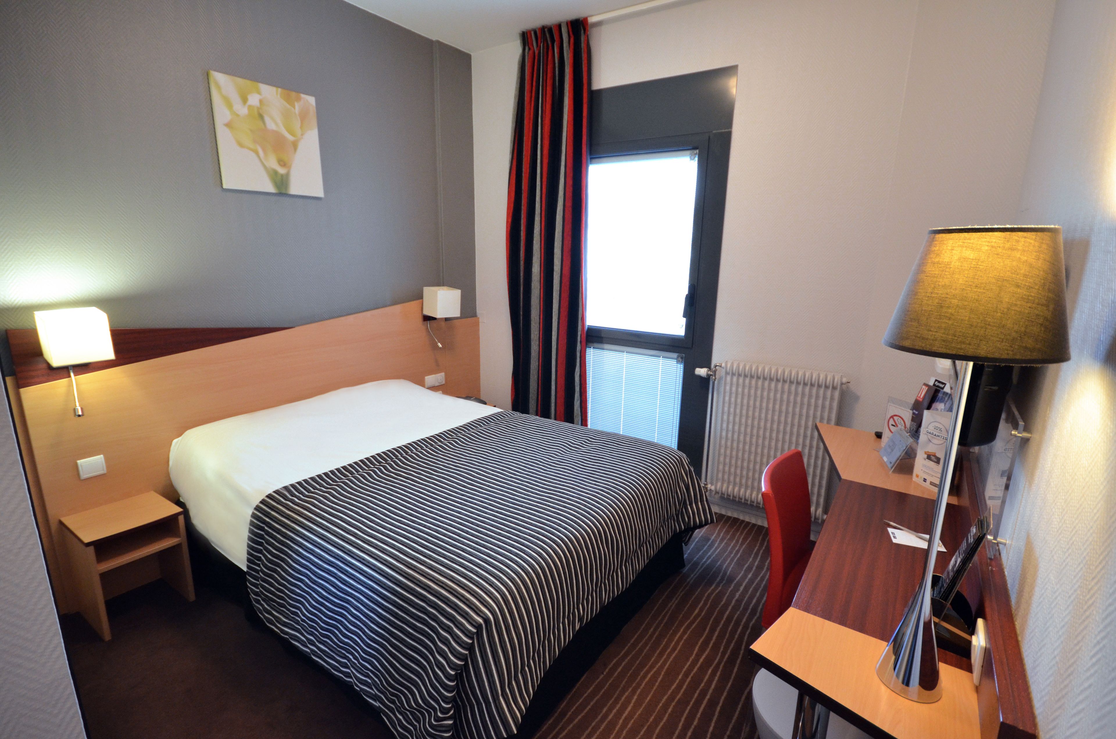 Habitación de hotel con cama, escritorio, silla, lámpara, ventana con cortinas, radiador y una obra de arte colgada en la pared.