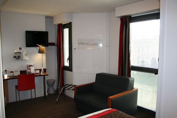 Habitación de hotel con escritorio, una silla, sofá, televisor y ventana con cortinas.