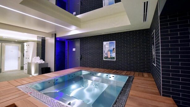 Un área de piscina interior moderna con cerramiento de vidrio, suelo de madera y pared de ladrillo negro.