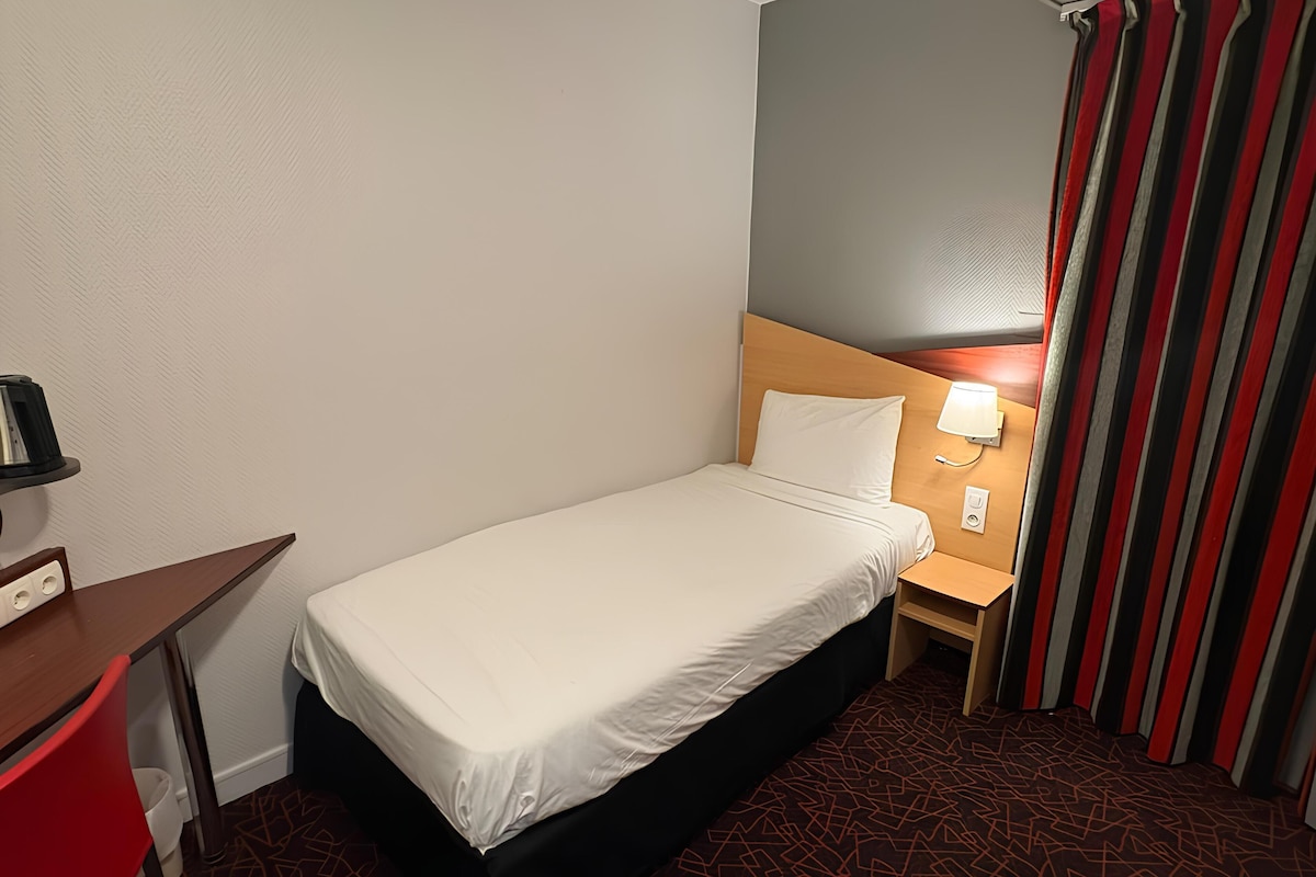Een kleine hotelkamer met een eenpersoonsbed, een nachtkastje met een lamp, een bureau met een stoel en een gestreept gordijn.