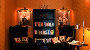 A room with two black armchairs, a bookshelf with books, framed pictures, and a small table with a lamp.