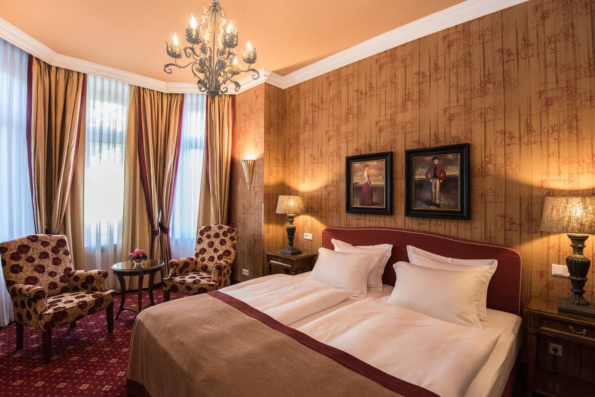 A hotel room with a large bed, two armchairs, a small table with a vase, a chandelier, and two wall-mounted lamps.