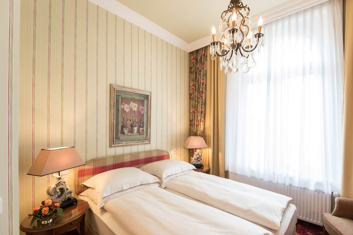 A hotel room with striped wallpaper, two beds, a chandelier, a painting, and a window with curtains.