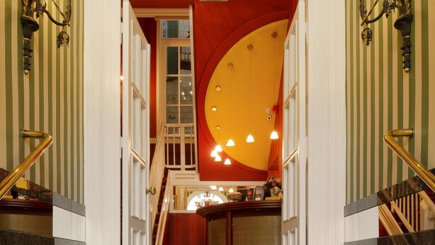 A grand staircase with a red and gold accent wall, leading to a second-floor area with a chandelier and wall sconces.