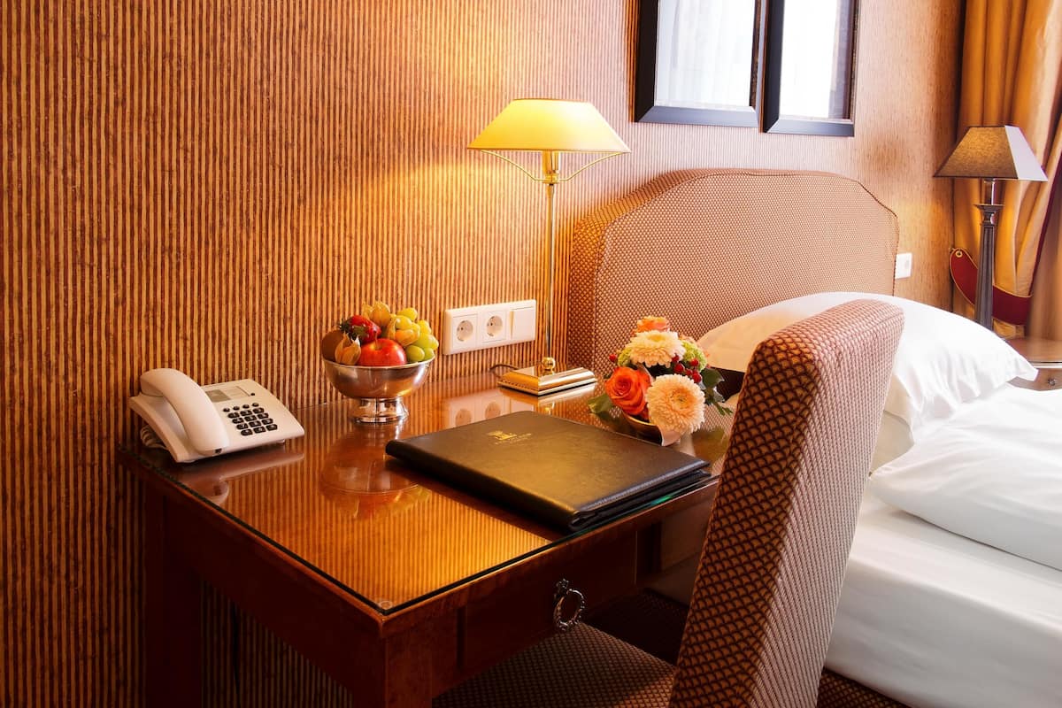 A hotel room with a wooden desk, a telephone, a bowl of fruit, a laptop, a flower vase, and a bed with white linens.